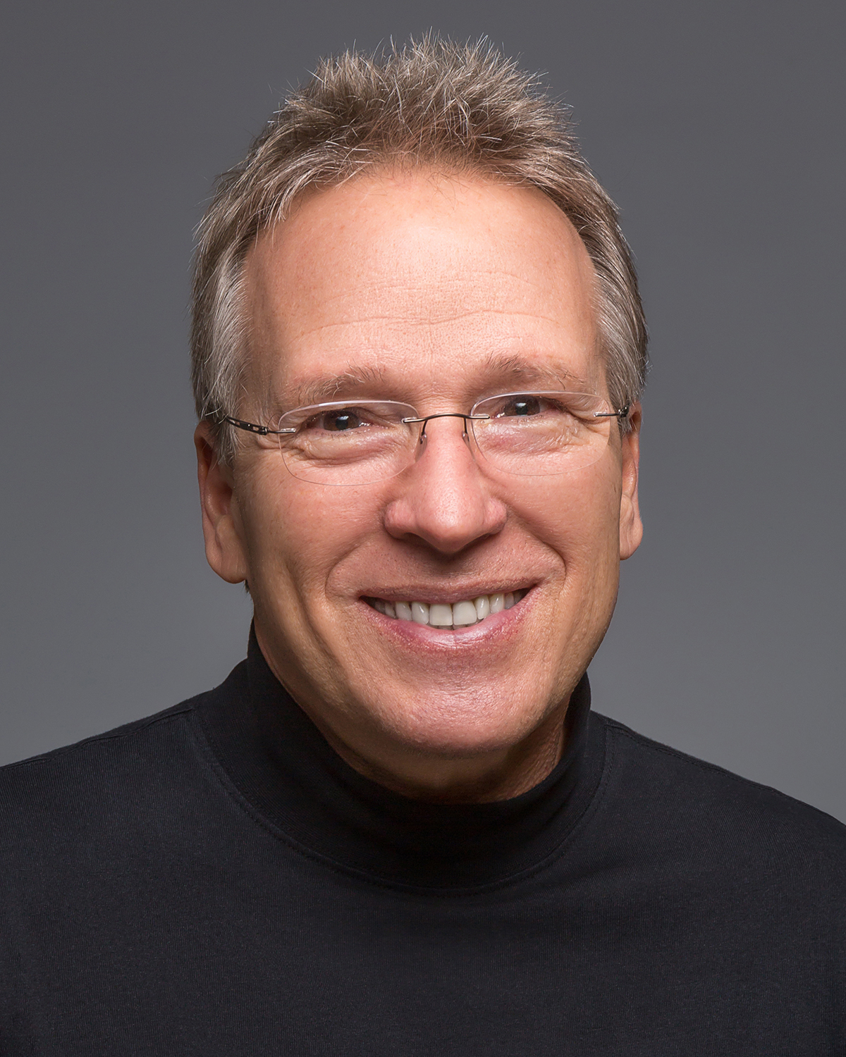Professional headshot of Dave Nelsen, smiling with a friendly demeanor. He's wearing a black turtleneck and rimless glasses, with short, sandy blond, graying hair. The background is a neutral gray, highlighting his approachable expression.